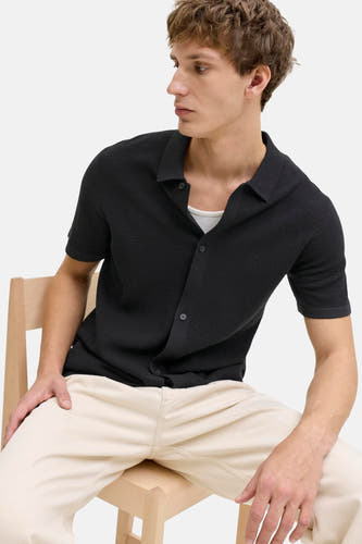 Pull noir de CORE BY JACK & JONES, à manches courtes et fermeture boutonnée, porté avec une chemise blanche et un pantalon beige.