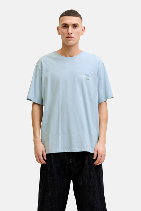 T-shirt bleu de ORIGINALS BY JACK & JONES avec imprimé discret sur la poitrine, porté avec un short foncé et des baskets blanches.