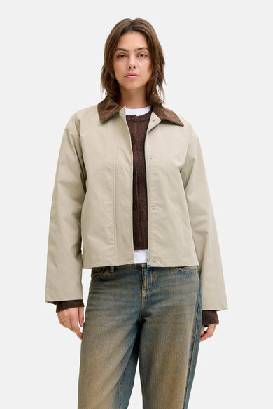 Manteau beige court de JJXX, avec un col marron et une fermeture éclair, porté sur un gilet marron et des jeans.