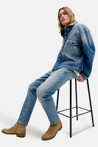 Jeans tapered mid blue denim van JACK & JONES JEANS INTELLIGENCE, gecombineerd met een denim jas en suède laarzen, zittend op een kruk.