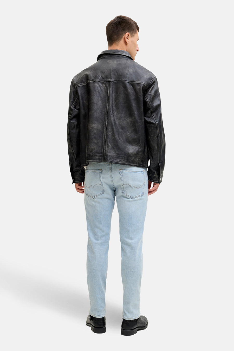 Man van achteren in leren jack, lichtblauwe jeans en zwarte schoenen.