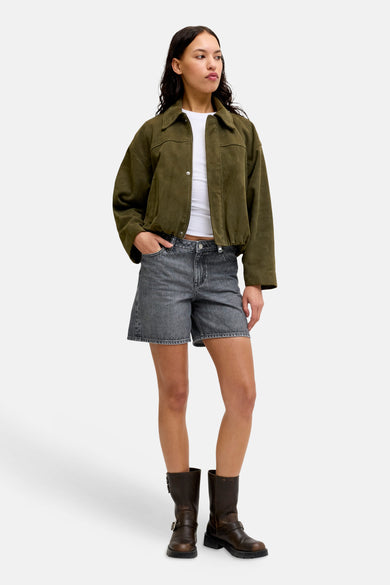 Donkergrijze denim short van JJXX, gedragen door een model met een olijfgroen jack, witte top en bruine laarzen.