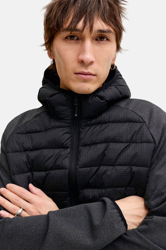 Portret van een man met een zwart en grijs gewatteerd jack met capuchon, gekruiste armen en ring.
