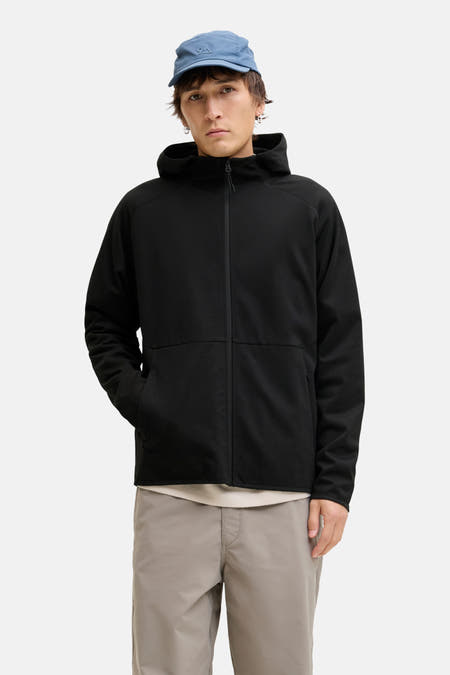 Zwarte sweater gilet van CORE BY JACK & JONES, met lange mouwen en een capuchon, gecombineerd met een blauwe pet en grijze broek.
