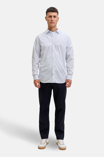 Een man staat rechtop in een lichtblauw overhemd, marineblauwe broek en witte sneakers.