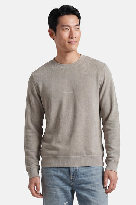 Pull gris à col rond de CORE BY JACK & JONES, avec une texture fine et un logo discret sur la poitrine, porté avec un jean clair.