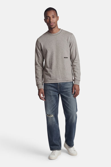 Pull gris à col rond de CORE BY JACK & JONES, avec logo noir 'JJCORE' sur la poitrine, porté par un homme avec un jean déchiré et des baskets blanches.