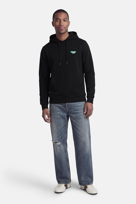Zwarte sweater met kap van CORE BY JACK & JONES, met blauw logo op de borst en gecombineerd met jeans met gaten en lage sneakers.
