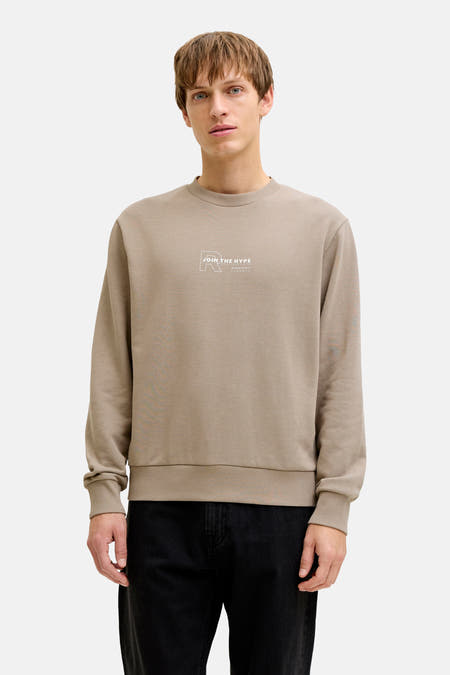 Bruine sweater met ronde hals van JJREBEL, met tekstprint op de borst, gedragen met een zwarte broek.
