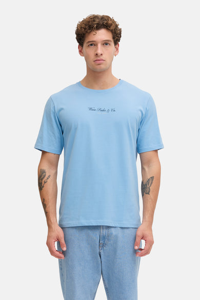 T-shirt bleu à manches courtes de JJREBEL, porté sous une chemise bleu marine avec un jean clair et des chaussures écru.
