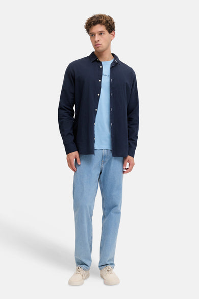 Blauw T-shirt met korte mouwen van JJREBEL, gedragen onder een marineblauw overhemd met lichte jeans en ecru schoenen.
