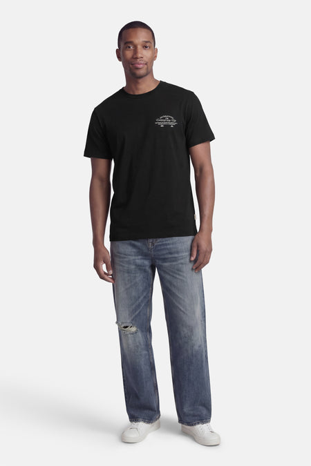 T-shirt noir à manches courtes de ORIGINALS BY JACK & JONES, avec imprimé blanc sur la poitrine ; porté avec un jean bleu et des baskets blanches.