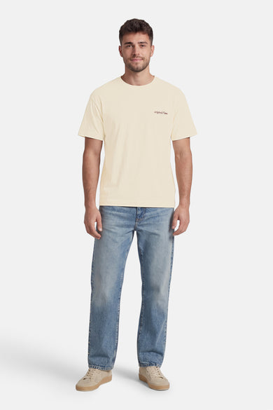 Beige T-shirt van ORIGINALS BY JACK & JONES met 'originals © 1990' print, gedragen door een man met een jeans met zijknopen en beige geperforeerde sneakers.