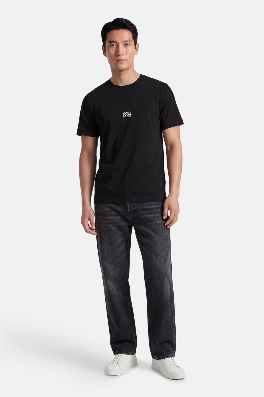 T-shirt noir de CORE BY JACK & JONES avec logo blanc sur la poitrine, porté par un homme avec un jean foncé et des baskets blanches.