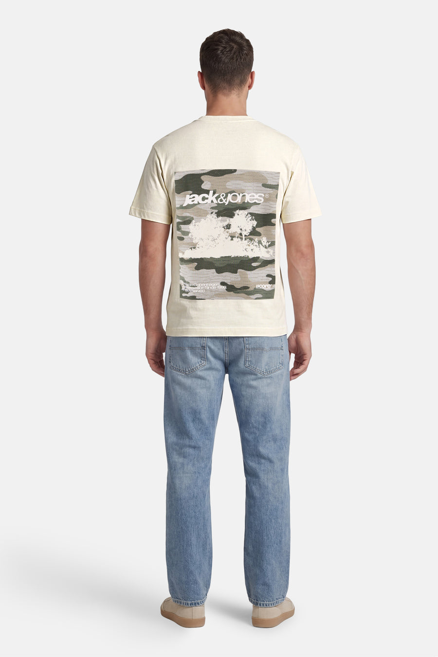 Ecru T-shirt van CORE BY JACK & JONES met camouflageprint en wit logo op de rug, gedragen door een man met blauwe spijkerbroek en beige schoenen (rugzicht).