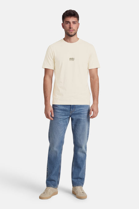 T-shirt écru de CORE BY JACK & JONES avec petit logo sur la poitrine, porté par un homme avec un jean bleu et des baskets beiges.