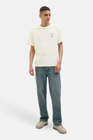 Een man in een crèmekleurig T-shirt met blauwe opdruk, blauwe jeans en beige schoenen, staand op een witte achtergrond.