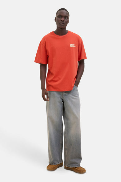 T-shirt orange à manches courtes d'ORIGINALS BY JACK & JONES, avec "Saint-Tropez" sur la poitrine, associé à des jeans larges gris et des mocassins bruns.