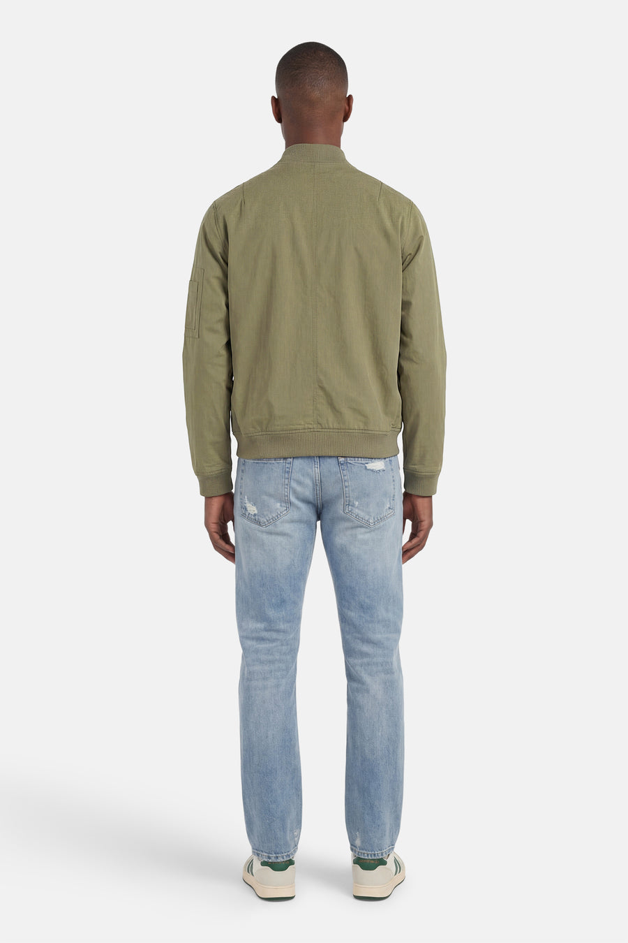 Veste courte verte de CORE BY JACK & JONES, vue de dos, avec texture tissée, poignets côtelés et poche sur la manche, portée avec un jean bleu clair et des baskets.