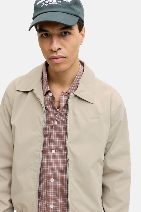 Manteau court beige de ORIGINALS BY JACK & JONES, avec fermeture éclair, porté sur une chemise à carreaux marron, avec casquette verte.