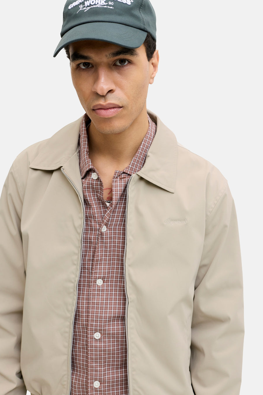 Manteau court beige de ORIGINALS BY JACK & JONES, avec fermeture éclair, porté sur une chemise à carreaux marron, avec casquette verte.