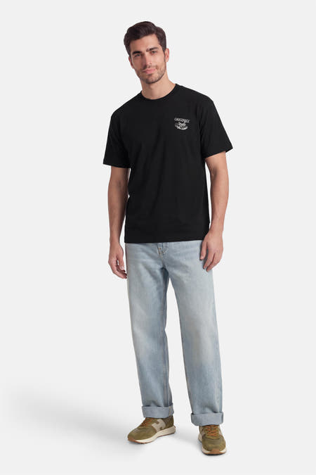 T-shirt noir à manches courtes de ORIGINALS BY JACK & JONES, avec un petit imprimé blanc sur la poitrine, porté avec un jean bleu clair et des baskets vertes.
