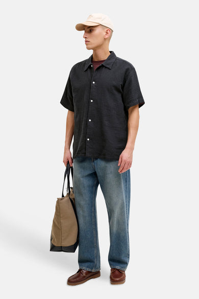 Chemise noire à manches courtes de ORIGINALS BY JACK & JONES, avec boutons blancs, portée avec un jean, des chaussures marron et une casquette beige.