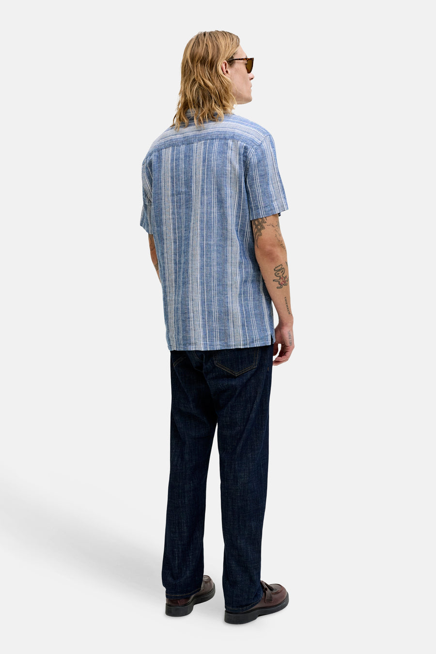 Blauw overhemd met korte mouwen van PREMIUM BLUE by JACK & JONES, gestreept, met tatoeages op de arm en donkere jeans.
