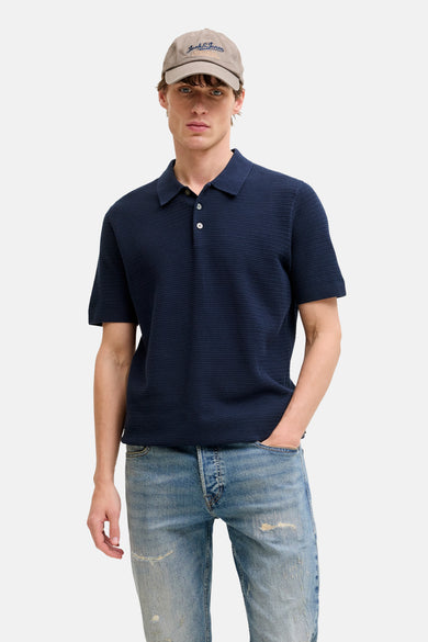 Blauwe polo met korte mouwen van PREMIUM BLUE by JACK & JONES, met een fijn weefsel, gedragen met een spijkerbroek en pet.

