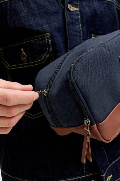 Blauwe toilettas van ACCESSORIES BY JACK & JONES, met bruin lederen accenten en ritssluiting, vastgehouden voor een donkerblauw denim jasje.