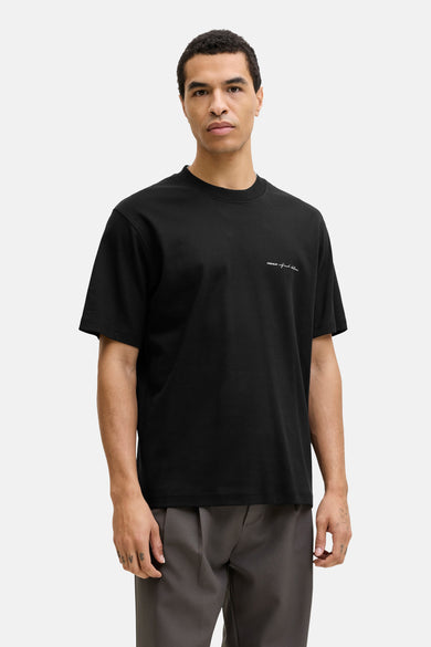 T-shirt noir à manches courtes de PREMIUM by JACK & JONES, avec un petit imprimé texte blanc et porté avec un pantalon gris.