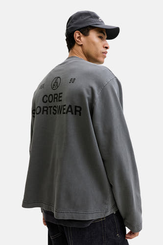 Zwarte sweater met ronde hals van CORE BY JACK & JONES, met print op de rug, gedragen met een donkere pet.
