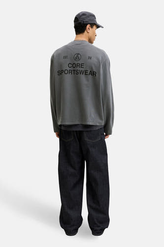 Zwarte sweater met ronde hals van CORE BY JACK & JONES, met logo op de rug, gedragen met een pet en wijde jeans.
