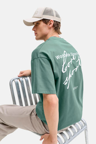 Groen T-shirt van CORE BY JACK & JONES met witte print op de rug, gedragen met een beige pet en broek.
