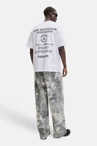 Wit T-shirt van CORE BY JACK & JONES met zwarte tekstprint op de rug, gedragen met grijze spijkerbroek.

