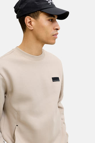 Beige sweater van CORE by JACK & JONES, met een zwarte logopatch op de borst en een zwarte pet.
