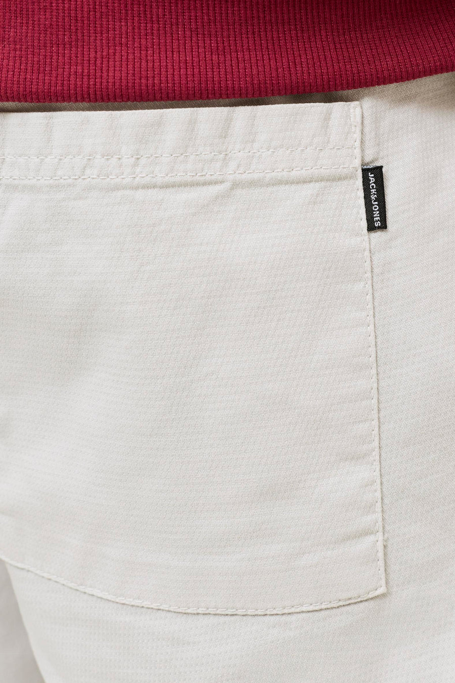 Beige short van JACK & JONES JEANS INTELLIGENCE met een geweven textuur, zichtbare zak met zwart label en een rood geribbelde stof erboven.