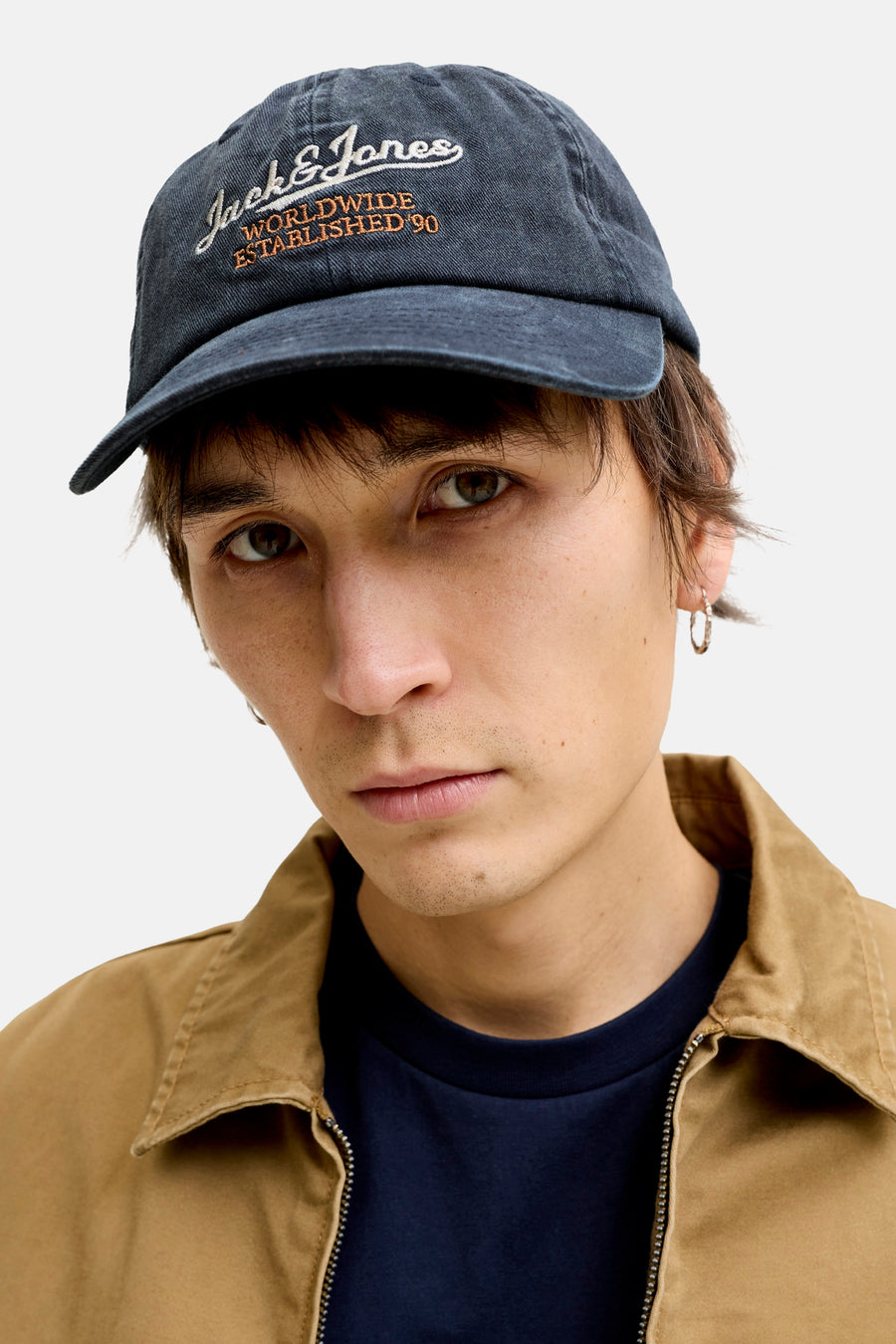 Blauwe pet van katoen met geborduurd logo van ACCESSORIES BY JACK & JONES, gedragen door model in close-up, beige jas en donkerblauw shirt zichtbaar