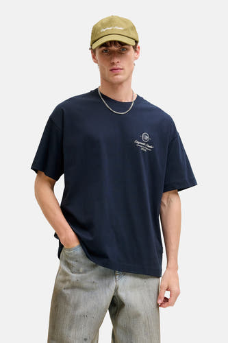 T-shirt bleu à manches courtes de Originals by Jack & Jones, avec un petit logo et porté avec une casquette, une chaîne, un jean et des bottes marron.