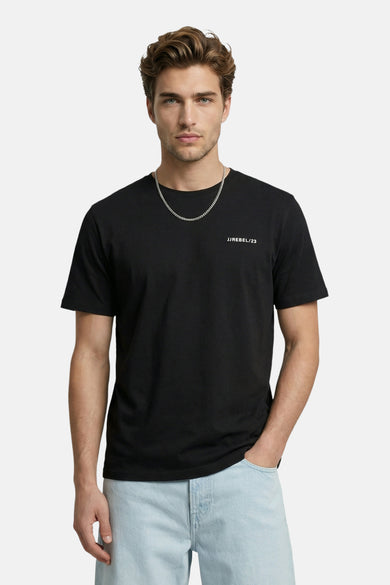T-shirt noir de JJREBEL avec imprimé blanc 'JJREBEL/23', porté par un homme avec une chaîne argentée et un jean bleu clair.
