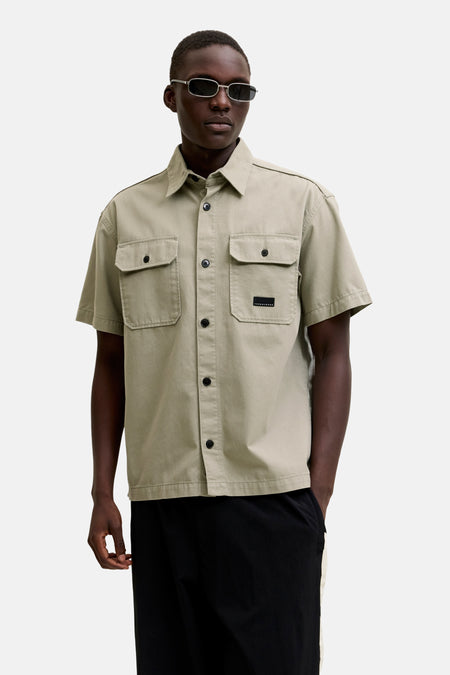Beige overhemd met korte mouwen van CORE BY JACK & JONES, met borstzakken en zwarte label, gedragen door een man met zonnebril en zwarte broek.