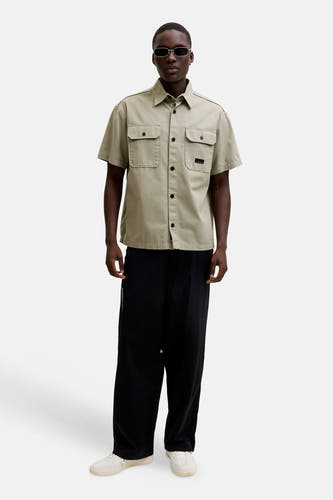 Chemise beige à manches courtes de CORE BY JACK & JONES, avec deux poches poitrine, boutons noirs et un écusson, portée avec un pantalon noir et des baskets blanches.