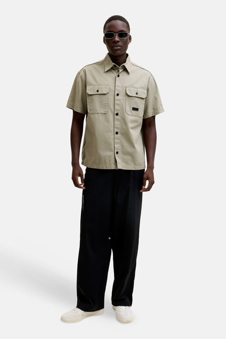 Beige hemd met korte mouwen van CORE BY JACK & JONES, met twee borstzakken, zwarte knopen en een patch, gedragen met een zwarte broek en witte sneakers.
