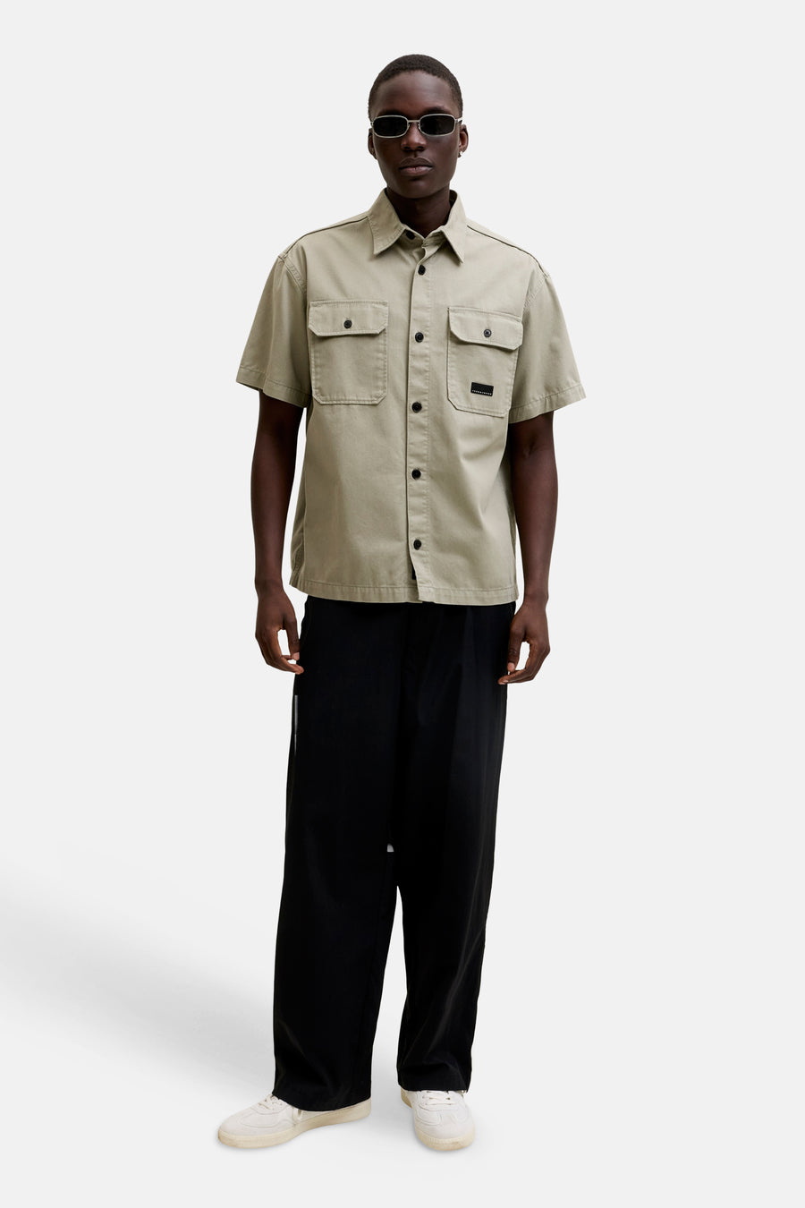 Chemise beige à manches courtes de CORE BY JACK & JONES, avec deux poches poitrine, boutons noirs et un écusson, portée avec un pantalon noir et des baskets blanches.