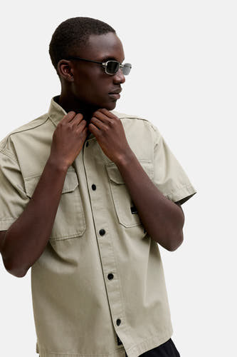 Chemise beige à manches courtes de CORE BY JACK & JONES, avec des poches poitrine de style utilitaire et des boutons noirs, portée par un mannequin avec des lunettes de soleil ajustant le col.