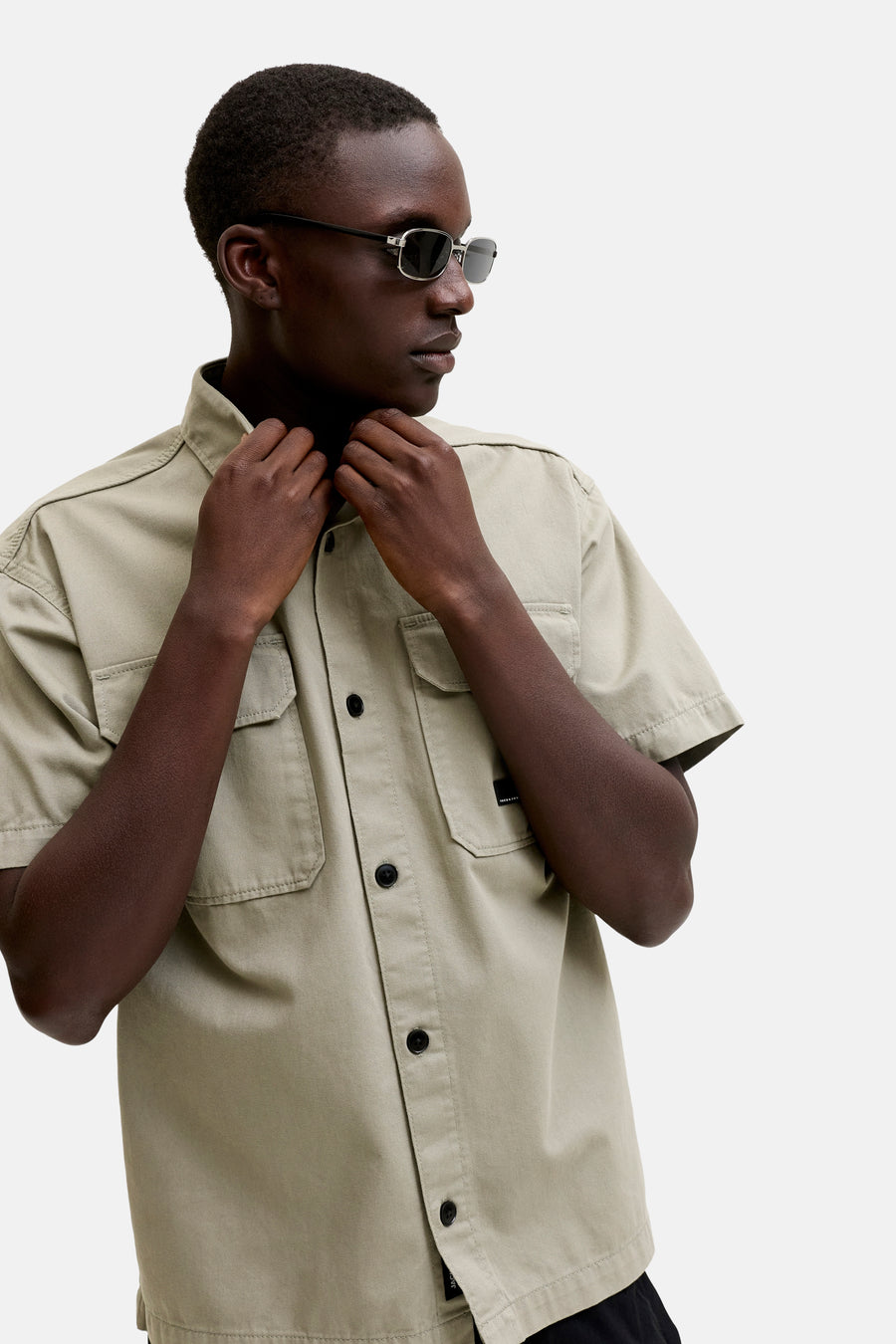 Chemise beige à manches courtes de CORE BY JACK & JONES, avec des poches poitrine de style utilitaire et des boutons noirs, portée par un mannequin avec des lunettes de soleil ajustant le col.
