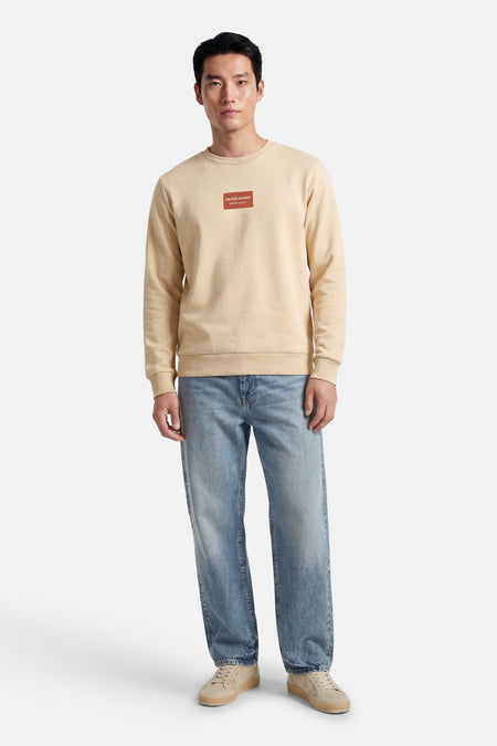 Beige sweater met ronde hals van CORE by JACK & JONES, met een rood logo op de borst, gecombineerd met blauwe jeans en beige schoenen.
