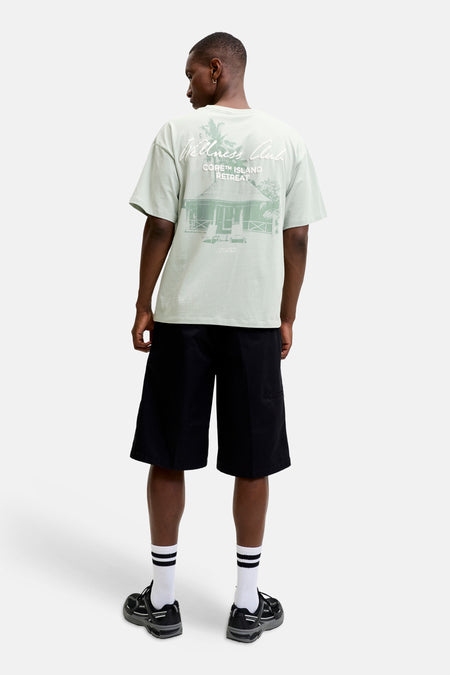 Groen T-shirt met korte mouwen van CORE BY JACK & JONES, met 'Wellness Club' en 'CORE™ ISLAND RETREAT' print op de rug, gedragen door een man met zwarte shorts en sneakers.