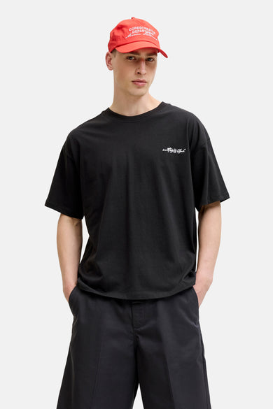 Zwart T-shirt met korte mouwen van CORE BY JACK & JONES, met witte 'wet 1999 Club' tekst op de borst, gedragen door een man met rode pet en zwarte korte broek.
