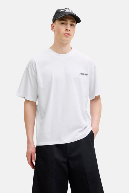 T-shirt blanc à manches courtes de CORE BY JACK & JONES, avec imprimé 'CORE 1980' sur la poitrine. Le mannequin porte également une casquette noire, un short noir et des chaussettes rayées.